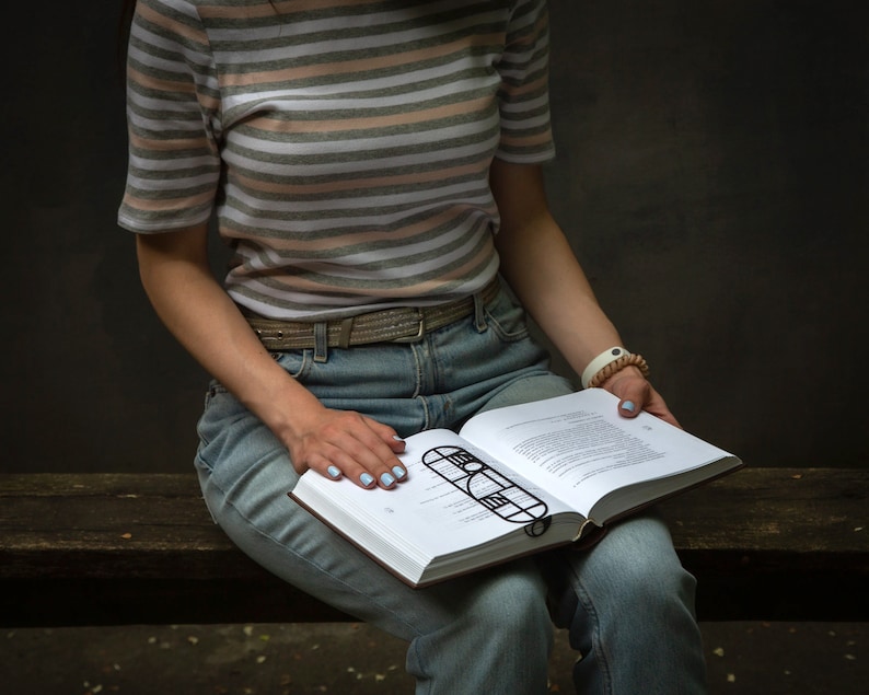Woman sitting on a bench reading a book with a black geometric Bauhaus-style metal bookmark placed across the pages — stylish and modern reading accessory.