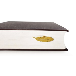 May include: A brown hardcover book with a gold feather bookmark. The bookmark is shaped like a feather and is placed on the top of the book.
