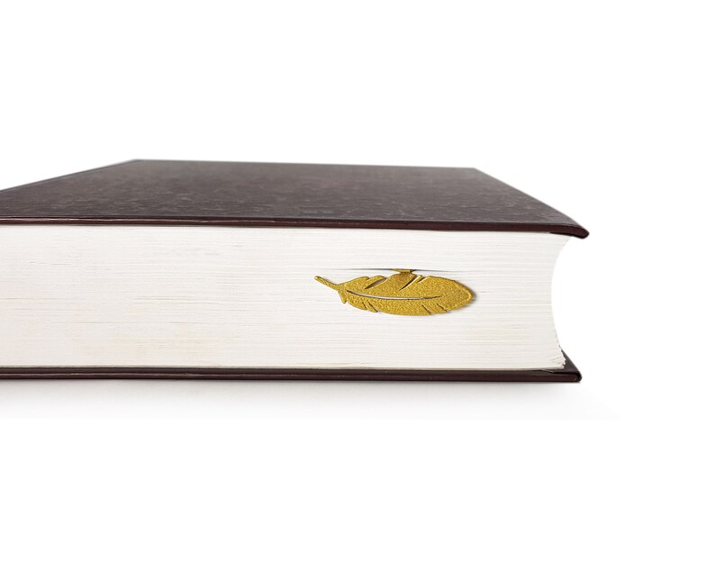 May include: A brown hardcover book with a gold feather bookmark. The bookmark is shaped like a feather and is placed on the top of the book.