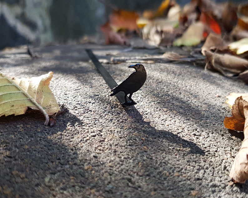 Black metal raven-shaped page saver photographed outdoors among autumn leaves and warm sunlight. A poetic, nature-inspired gift for book lovers with a dark, literary soul.