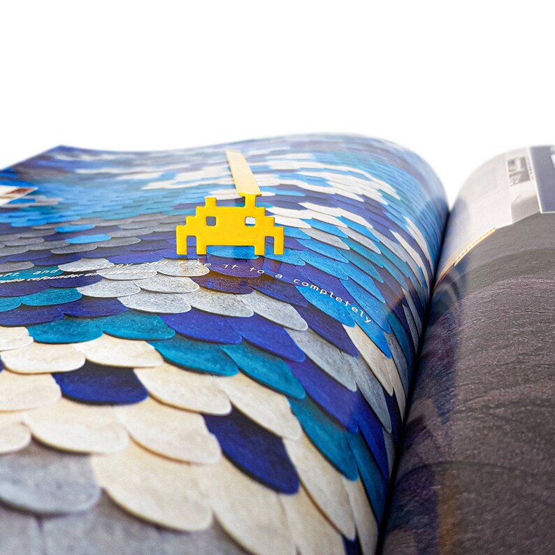 May include: A yellow plastic bookmark shaped like a retro video game character, a space invader, is placed on a page of a book with a blue and white patterned cover. The text "completely" is visible on the page.