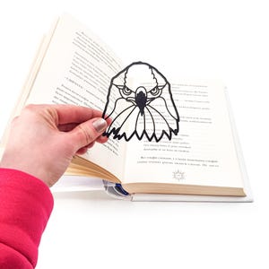 Op de afbeelding: Een zwarte papieren uitsnijding in de vorm van een adelaarskop. De adelaar heeft een scherpe snavel en intense ogen. De bladwijzer is in een boek geplaatst.