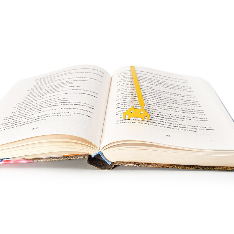 May include: A yellow metal bookmark shaped like a spaceship is placed in an open book. The book is open to page 119.