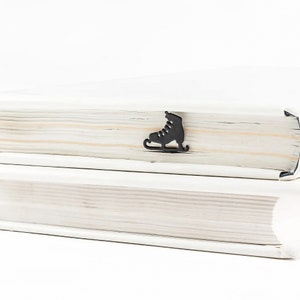 Minimalist-style metal bookmark Ice Skates. A metal stick is resting between the pages of the book. An ice skate is on the side of the book. It is visible even when the book is closed. Available in black or pink color.