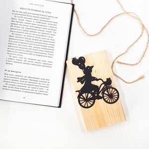 Black metal bike bookmark shaped as a girl riding a bicycle with balloons, placed on a wooden engraved block next to an open book on a white background.