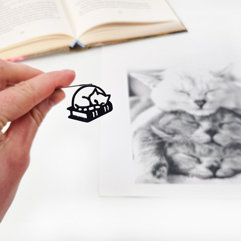 Black metal bookmark - Cat sleeping on the book. A long flat metal stick stays between the pages of the book. The sleeping on the book kitty is on the side of the book. It is visible even when the book is closed. Made by us in Ukraine.