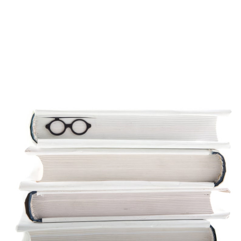 Architect-inspired black metal page saver shaped like Bauhaus-style round glasses, subtly tucked into a stacked set of white hardcovers. A minimalist tribute to modern design and reading culture
