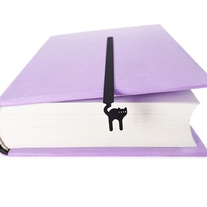 May include: A lavender book with a black cat-shaped bookmark. The bookmark is inserted between the pages and features a silhouette of a cat. The book has a textured cover and white pages.