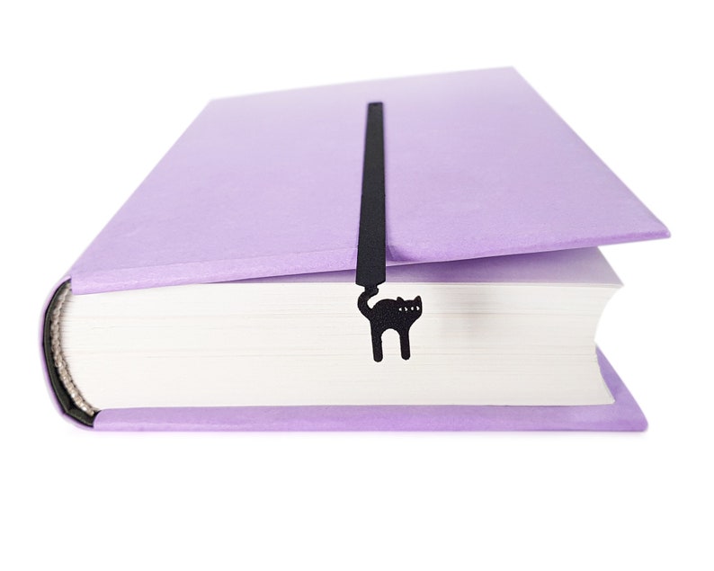 May include: A lavender book with a black cat-shaped bookmark. The bookmark is inserted between the pages and features a silhouette of a cat. The book has a textured cover and white pages.