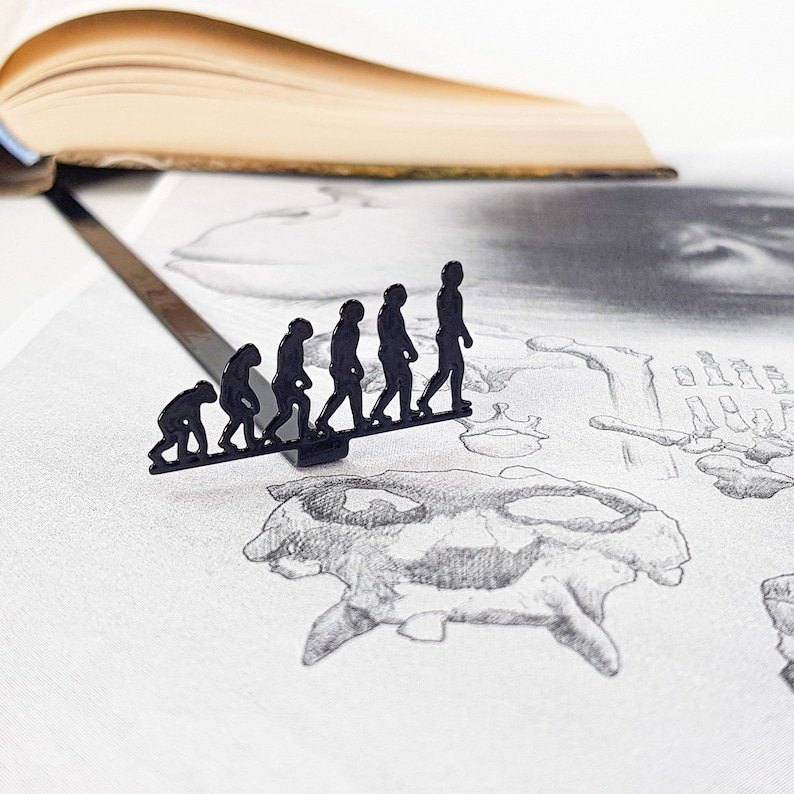 May include: A black metal bookmark depicting a line of human figures in various stages of evolution. The bookmark is on a white surface with a pencil drawing of a skull and bones.