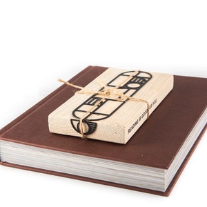 Black Bauhaus-style metal bookmark tied to an eco-friendly wooden block, resting on a closed brown hardcover book — minimalist gift presentation. The block has the text 'Reading is good for you' printed on it.