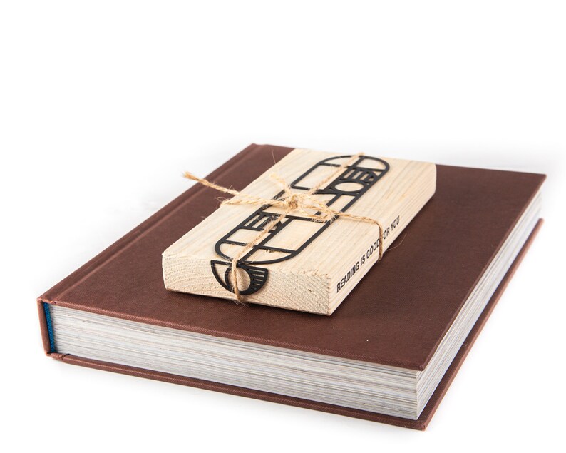 Black Bauhaus-style metal bookmark tied to an eco-friendly wooden block, resting on a closed brown hardcover book — minimalist gift presentation. The block has the text 'Reading is good for you' printed on it.