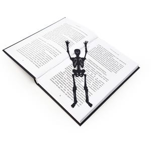 A black metal skeleton-shaped bookmark lying across the center of an open book, posed as if climbing up the pages.