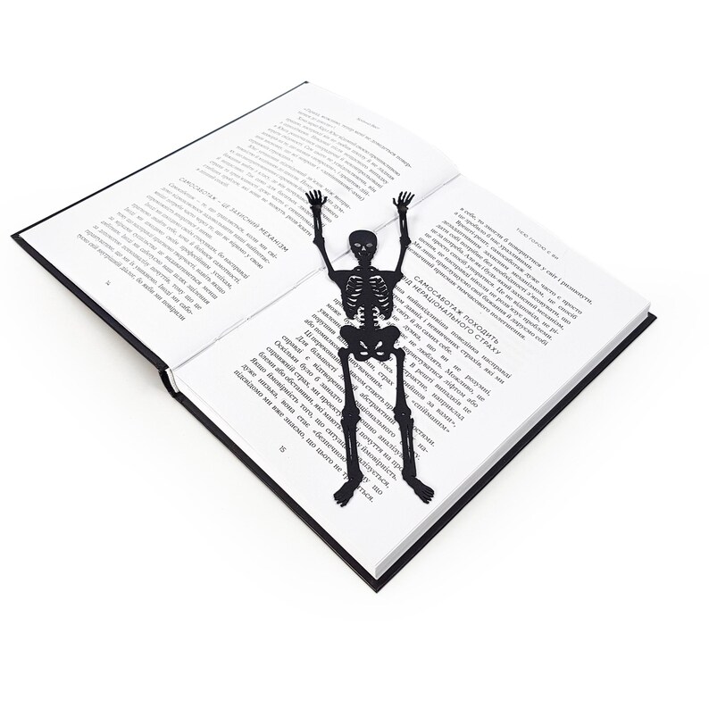 A black metal skeleton-shaped bookmark lying across the center of an open book, posed as if climbing up the pages.