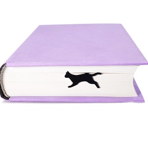 Black metal running cat bookmark peeking out from the closed pages of a lavender-colored hardcover book.
