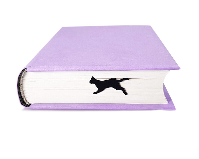 Black metal running cat bookmark peeking out from the closed pages of a lavender-colored hardcover book.