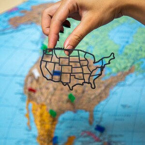 Può includere: Un contorno nero della mappa degli Stati Uniti viene tenuto su una mappa del mondo. La mappa è divisa in stati e include i nomi degli stati. La mappa è tenuta da una mano.