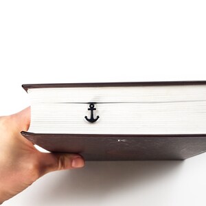 May include: A brown hardcover book with a black anchor bookmark. The book is being held by a hand.