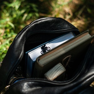 A black leather backpack with a zipper open, revealing a book, a small wooden block, and an elegant black metal bookmark with a figurine of a person riding a bicycle.
