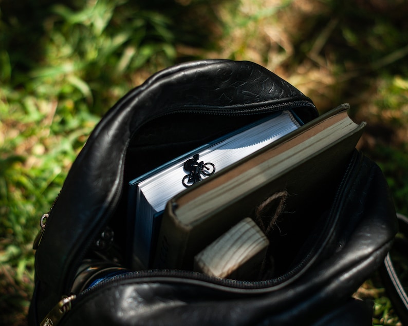 A black leather backpack with a zipper open, revealing a book, a small wooden block, and an elegant black metal bookmark with a figurine of a person riding a bicycle.