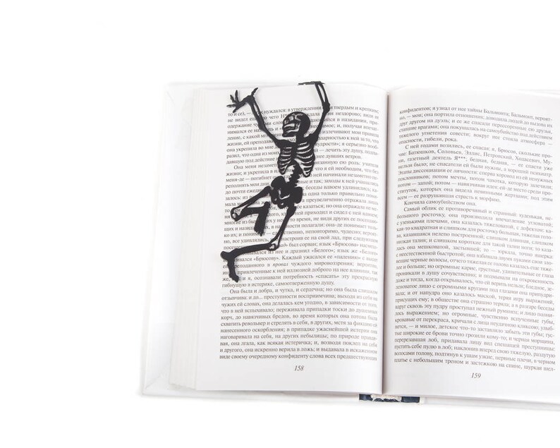 Bookmark Funny Dancing Skeleton by Coolbookmark. A large figure of the dancing skeleton stays between the pages of the book. The book in its hands in on the side of the book. It is visible even when the book is closed.