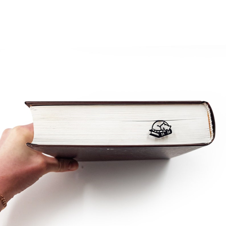Op de afbeelding: Zwarte metalen boekenlegger met een ontwerp van een slapende kat op twee boeken. De boekenlegger is geplaatst in een dik, bruin gebonden boek met witte pagina's. De afbeelding is tegen een witte achtergrond.