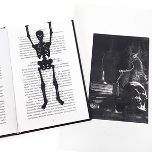 A black metal skeleton-shaped bookmark placed inside an open book, with a dark, atmospheric illustration showing a skull, candle, and old books beside it.