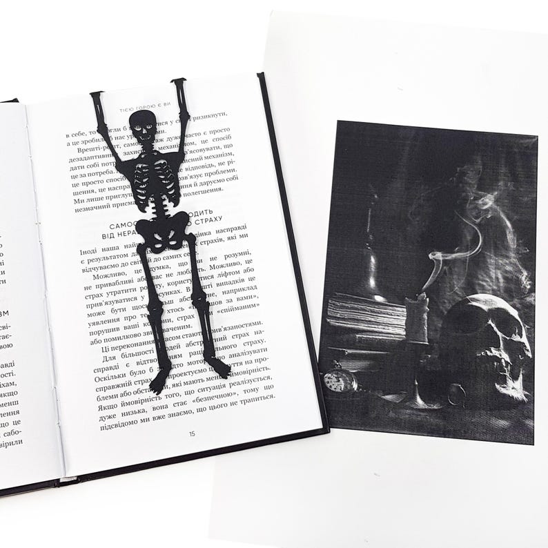 A black metal skeleton-shaped bookmark placed inside an open book, with a dark, atmospheric illustration showing a skull, candle, and old books beside it.
