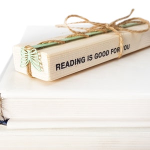 May include: A wooden bookmark with the text "READING IS GOOD FOR YOU" and a green butterfly design. The bookmark is tied with twine and is resting on top of two white books.