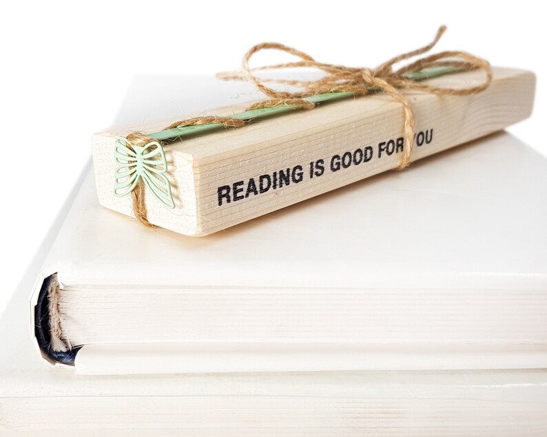 May include: A wooden bookmark with the text "READING IS GOOD FOR YOU" and a green butterfly design. The bookmark is tied with twine and is resting on top of two white books.
