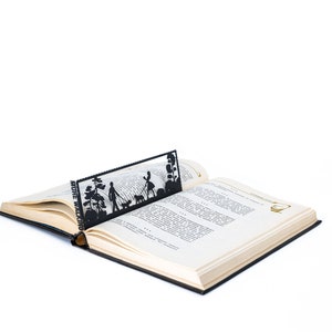 May include: A black metal bookmark with a silhouette of a couple walking a dog in a park. The bookmark is placed in an open book with the text "MORE WALKING" visible.