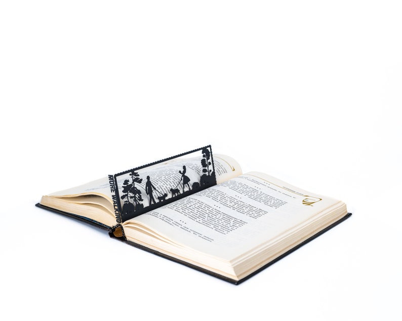 May include: A black metal bookmark with a silhouette of a couple walking a dog in a park. The bookmark is placed in an open book with the text "MORE WALKING" visible.