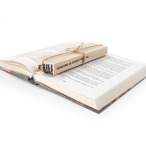 May include: A wooden bookmark with the text "Reading is good for you" and a silhouette of a group of people walking. The bookmark is placed on top of an open book.