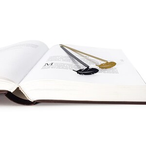 May include: Two feather-shaped bookmarks, one gold and one black, are placed in a book. The book is open and the pages are white.