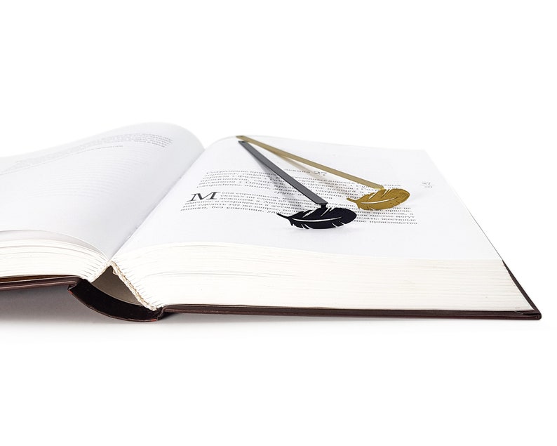 May include: Two feather-shaped bookmarks, one gold and one black, are placed in a book. The book is open and the pages are white.
