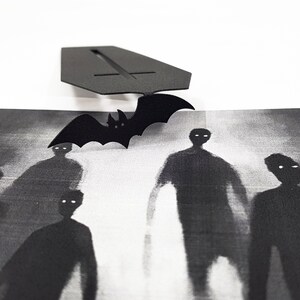 Black metal coffin-shaped bookmark with a flying bat detail placed above a monochrome artwork showing ghostly, shadowy figures with glowing eyes.