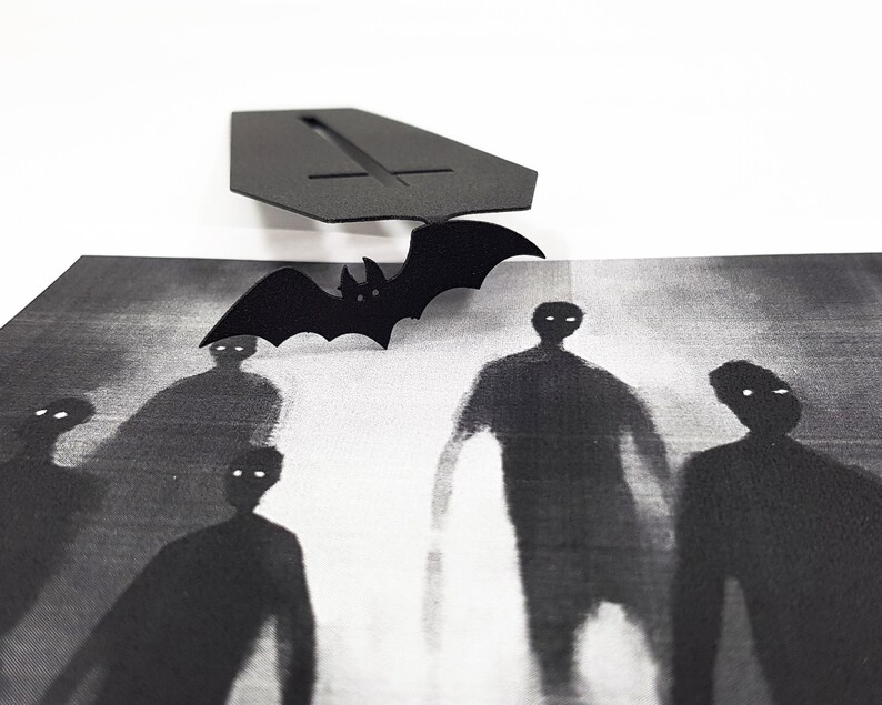 Black metal coffin-shaped bookmark with a flying bat detail placed above a monochrome artwork showing ghostly, shadowy figures with glowing eyes.
