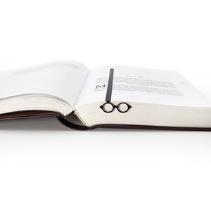 Black metal page saver in the shape of iconic architect glasses, placed in the center of an open book. Sleek, Bauhaus-inspired design for modern readers.