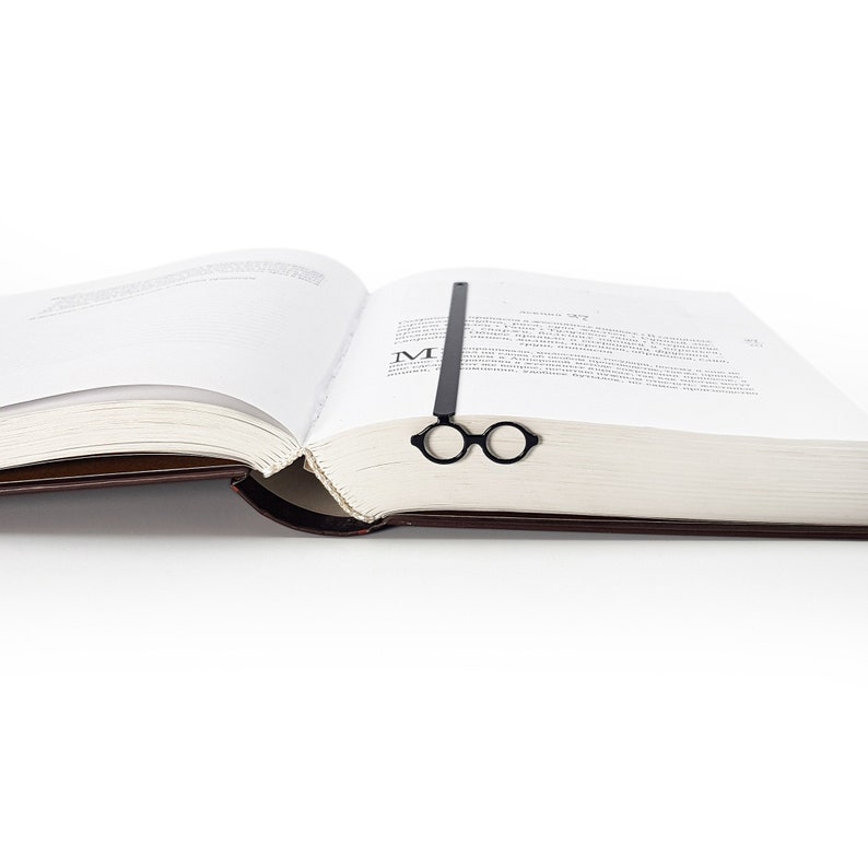 Black metal page saver in the shape of iconic architect glasses, placed in the center of an open book. Sleek, Bauhaus-inspired design for modern readers.