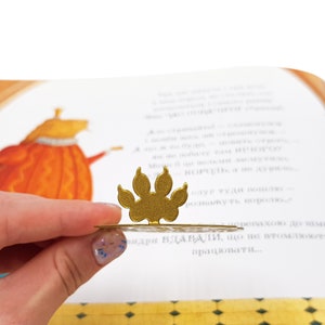 May include: A gold paw print bookmark with a decorative design. The bookmark is resting on a page of a children's book.