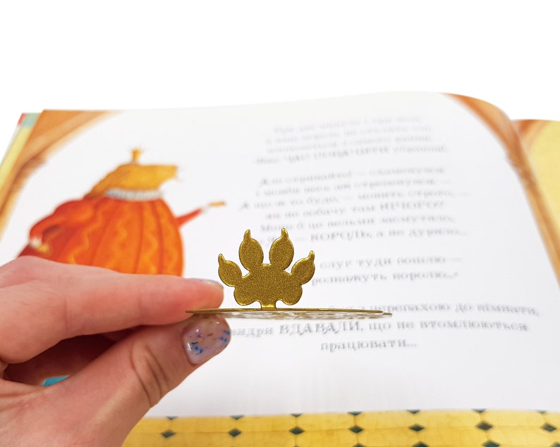 May include: A gold paw print bookmark with a decorative design. The bookmark is resting on a page of a children's book.