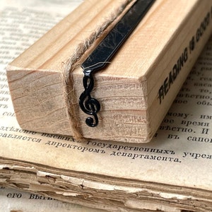 May include: A wooden bookmark with a black metal treble clef charm and the text "Reading is good for you" stamped on the side. The bookmark is resting on a stack of old books.