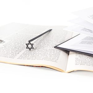 Black Star of David metal bookmark lying across an open book with English text, surrounded by scattered pages and books, minimalist Judaica-inspired design.