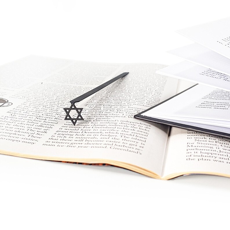 Black Star of David metal bookmark lying across an open book with English text, surrounded by scattered pages and books, minimalist Judaica-inspired design.