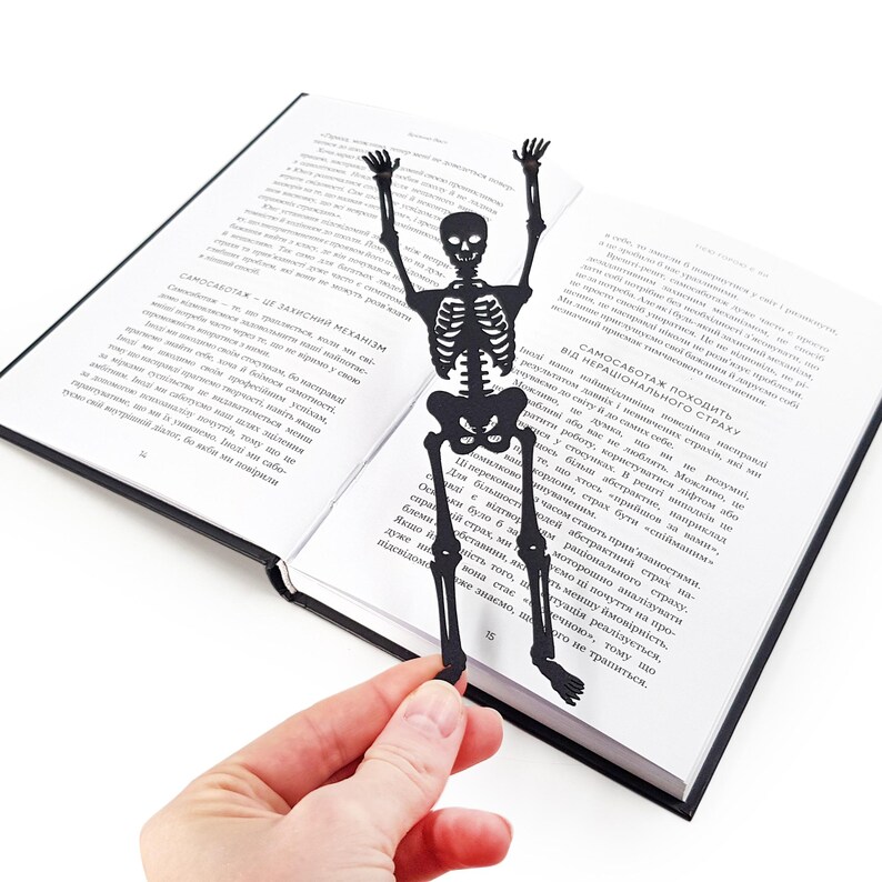 A hand holding a black metal skeleton-shaped bookmark on top of an open book, the skeleton positioned as if climbing out from the center of the pages.