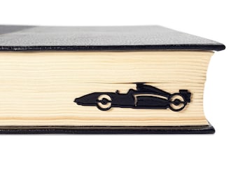 May include: A black race car bookmark is inserted into the pages of a book. The bookmark is made of metal and is in the shape of a Formula 1 race car. The book has a black cover and light-colored pages.