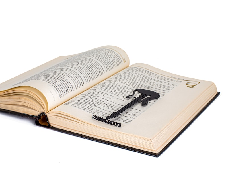 Black metal bookmark Reading Rocks. The cut out of metal silhuette of the guitear is sitting between the pages of the book. The writing Reading Roack is seen on the side of the book even when the book is closed. Designed and made by Coolbookmark on Etsy.