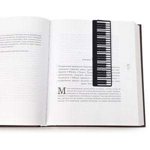 Black metal bookmark Piano Player. The line of piano keys stays between the pages of the book. A silhouette of the lady in a long dress playing the piano stays on the side of the book even when the book is closed. Made by us in our workshop.