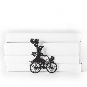 Black metal bike bookmark shaped as a girl riding a bicycle with balloons, leaning against a stack of white books on a white background.
