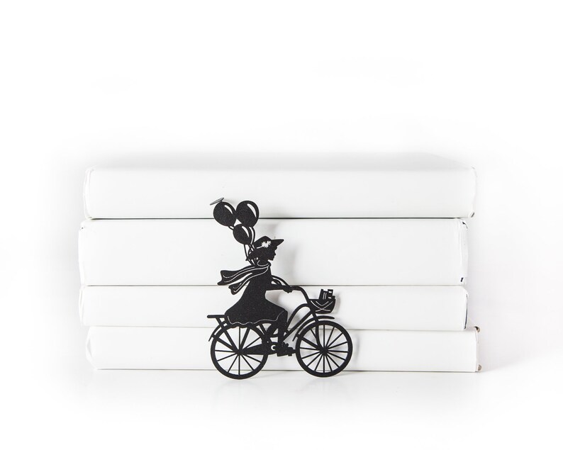 Black metal bike bookmark shaped as a girl riding a bicycle with balloons, leaning against a stack of white books on a white background.
